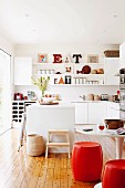 weiße Küche mit Küchenblock und Dielenfussboden; im Vordergrund rote fassförmige Sitzhocker an rundem Tisch, im Hintergrund Einbauelement mit Wandboard und Dekoration