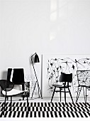 Schwarze Stühle aus verschiedenen Epochen auf schwarz-weiss gestreiftem Teppich vor Zeichnungen, die an Wand lehnen
