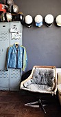 Vintage swivel chair in front of motorbike jacket hanging on locker and collection of helmets on grey wall