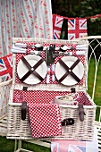 Plates and cutlery in the lid of an open, white picnic basket lined with red and white spotted fabric and stylised Union Jack bunting in the background
