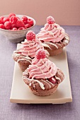 Three cupcakes with raspberry cream topping, with a bowl of raspberries in the background