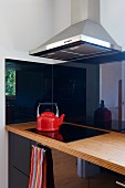 Corner of modern kitchen with reflective, black cupboard doors and red kettle on black ceramic hob