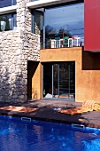 Modern house with large windows and stone walls; azure blue swimming pool with wooden terrace in foreground