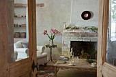View through rustic double doors of simple coffee table in front of masonry fireplace, bouquet of lilies on bistro table and country-style armchair