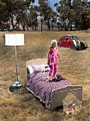 Girl bouncing on bed next to lit standard lamp in twilight meadow with old Beetle in background