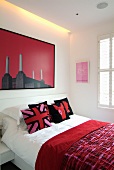Red patterned bedspread on bed and modern picture on white wall with indirect lighting