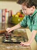 A little boy spooning jam into tarts in a muffin tin