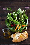 Spring vegetables in a green pot with white bread and a small jug of olive oil