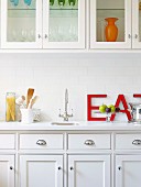 Elegant, white, country-house-style fitted kitchen with wall units; large red letters leaning on wall