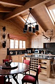 Round dining table and upholstered chairs in front of island counter and open-plan kitchen in attic storey of wooden house