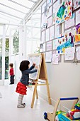 Kleines Mädchen an einer Maltafel und Sammlung angepinnter Kinderzeichnungen in weißem Raum mit Glasdach