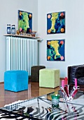 Glass coffee table on zebra skin rug, brightly coloured cubic stools in front of radiator and series of three pop art-style pictures of cows' heads