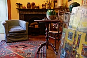 Corner of vintage-style living room - armchair with loose cover in front of open fireplace and wooden side table next to stack of paintings