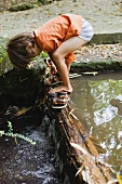 Junge zieht seine Sandalen an auf einem Baumstamm über einem Bach