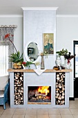 Offener Kamin mit Brennholzfächern und Kaminsims mit Aloe und Gartenpflanzen, darüber Wandspiegel und Blumenbild