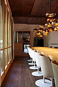 Long table and white shell chairs next to glazed facade below decorative arrangement of gold-coloured pendant lamps (Schloss Schauenstein)