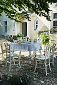 Table set with white cloth and crockery and wooden chairs on gravel surface in front of old country house