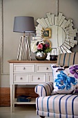 Lamp with lampshade and vase of flowers on cabinet in front of sun-shaped mirror