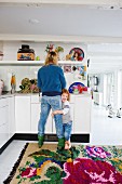 Mutter bei der Küchenarbeit mit Kind am Bein; weiße Raumflucht mit farbenfrohem Teppich auf dem Boden