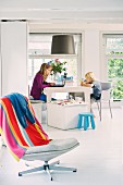 Two children with laptop and toys sitting in cool, white dining area; colourful striped towel on swivel chair in foreground