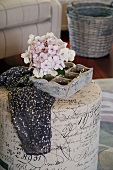 Holzkästchen mit Hortensienblüten und schwarzer Stoff mit Pailletten auf einem zylinderförmigen Polsterhocker