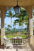 White rattan chairs at set table on colonial-style veranda with view into garden of palm trees