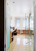 Blick in Badezimmer mit beigen Bodenfliesen; Wäschekörben unter Steinwaschtisch und halboffene Duschtür