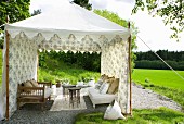 Benches and delicate side tables in tent-like pavilion of floral fabric in green landscape