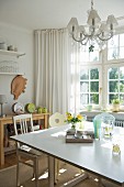 Sunny, simple kitchen with bay window, square dining table and vintage chairs; crystal chandelier above table