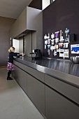 Girl at long counter of designer kitchen made from grey HPL and stainless steel; mementos pinned to wall