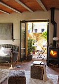 Sitzwürfel mit Fellbezug auf Teppich und Kanonenofen mit Feuer neben offenen Terrassentüren im Wohnzimmer eines Landhauses