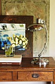Retro Tischleuchte mit Chromgestell neben Blumenstrauss auf schlichtem Wandtisch vor gerahmtem Bild mit Architekturmotiv
