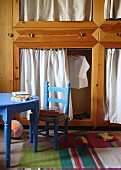 Blauer Kinderstuhl und blauer Tisch vor Holzschrank mit Vorhängen in den Öffnungen
