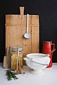 Vintage soup tureen and pasta in storage jar in front of chopping board leaning on black wall