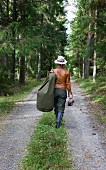Woman wearing smart, traditional country-style clothing walking along woodland path with duffle bag over shoulder