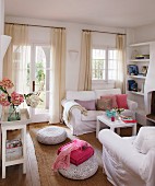 White living room furniture and floor pillows on a sisal rug in a living room with airy curtains at glazed doors and windows