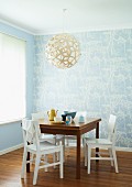 Breakfast table and white, wooden chairs beneath a round, wicker hanging lamp in the corner with bright blue wall paper with a white floral pattern