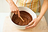 A woman stirs chocolate cake mixture with a wooden spoon in a bowl