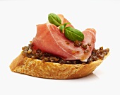 A slice of baguette topped with olive purée, Parma ham and basil leaves