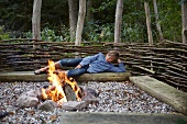 Boy lying on log bench next to bonfire