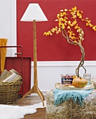 Tray of groceries on stool in front of standard lamp with white lampshade and wooden base and small tree against red-painted wall