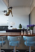 Mythical figure hanging from ceiling and bar stools with light blue upholstery at counter in open-plan loft apartment kitchen