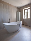 Free-standing designer bathtub and standpipe tap fitting in modern bathroom with pale tiles on walls and floor