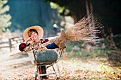 Woman with besom broom sitting in wheelbarrow of autumn leaves in garden