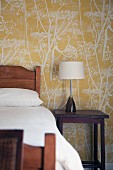 Holzbett und Lampe auf Nachttisch vor Tapete mit floraler Muster