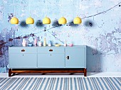 Row of yellow pendant lamps above sideboard painted pale blue on wooden base frame in front of wall with crumbling plaster