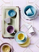 Top view of cups in pastel shades, some on dishes and saucers on patterned, retro tablecloth