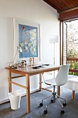 Modern swivel office chair at wooden table below picture on wall next to standard lamp and open balcony door