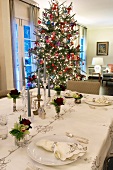 Dezent gemusterte Tischdecke mit Servietten und Rosendeko auf festlicher Tafel mit Weihnachtsbaum im Hintergrund