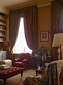 Comfortable armchairs with loose covers, ottoman and draped curtains with tassels in high-ceilinged salon of historic country house